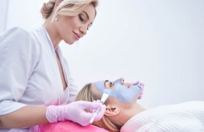 Cosmetician spreading blue clay paste over facial skin of client using two brushes